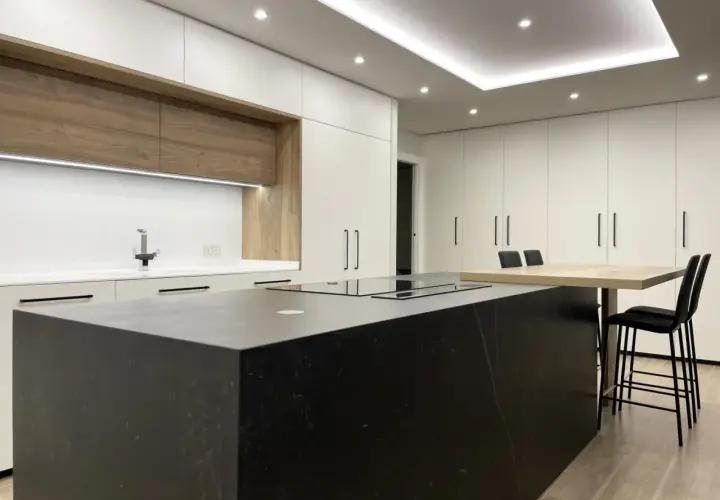 Cocina moderna con isla negra, encimeras blancas y armarios de madera. La iluminación LED bajo los armarios y en el techo resalta el diseño, mostrando cómo iluminar una cocina con estilo.