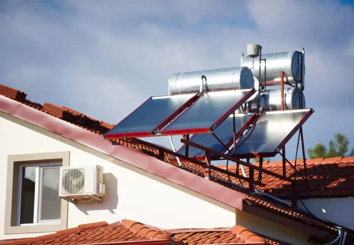 Paneles solares y tanques para agua caliente solar en un tejado de tejas rojas, bajo un cielo azul con nubes.