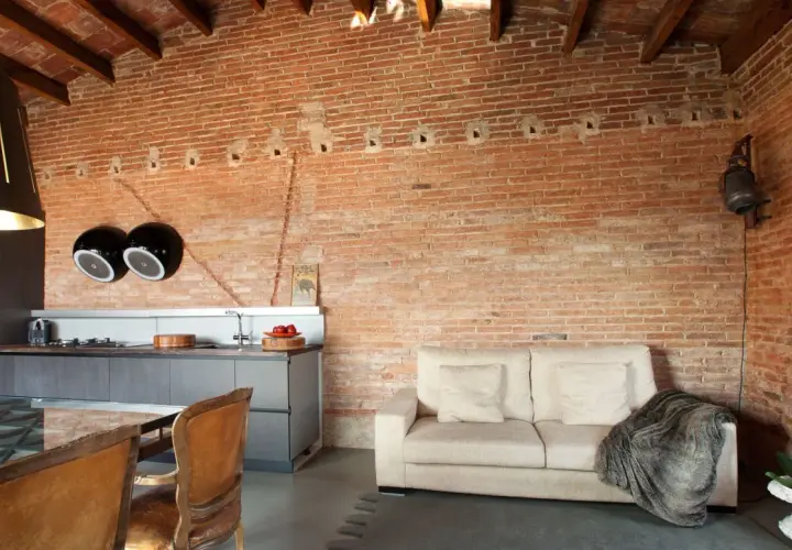 Un salón moderno con una pared rústica interior de ladrillo visto, sofá beige, cocina integrada y sillas de cuero.