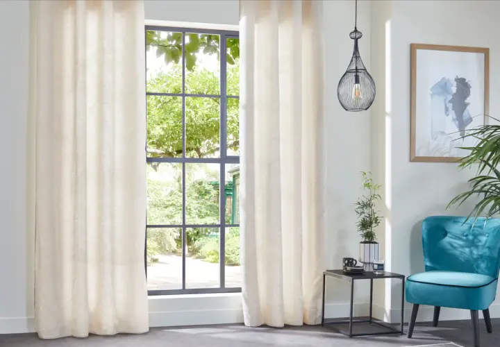 Elegantes cortinas de tela crema con sutil estampado, ideales para distintos tipos de cortinas para ventanas, complementan un sillón turquesa y un cuadro abstracto.