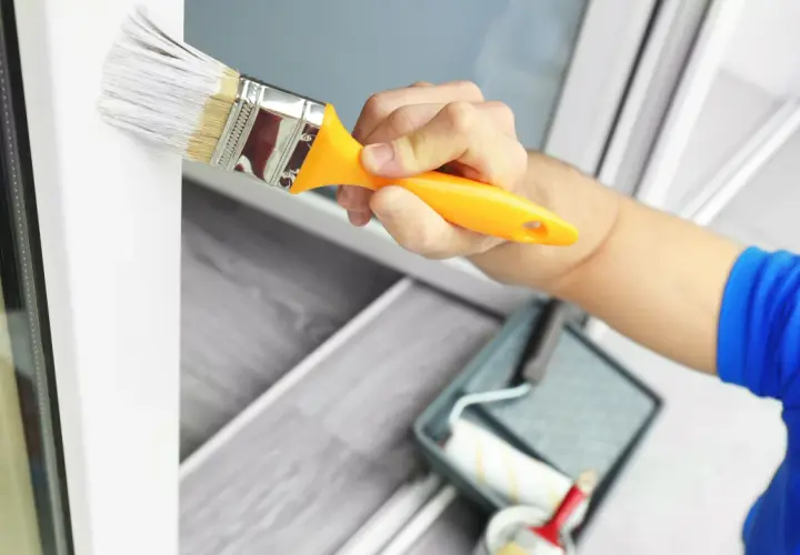 Mano pintando un marco de ventana blanco con una brocha. Se ve una bandeja de pintura y un rodillo en el fondo. Ideal para pintura para pintar plástico.