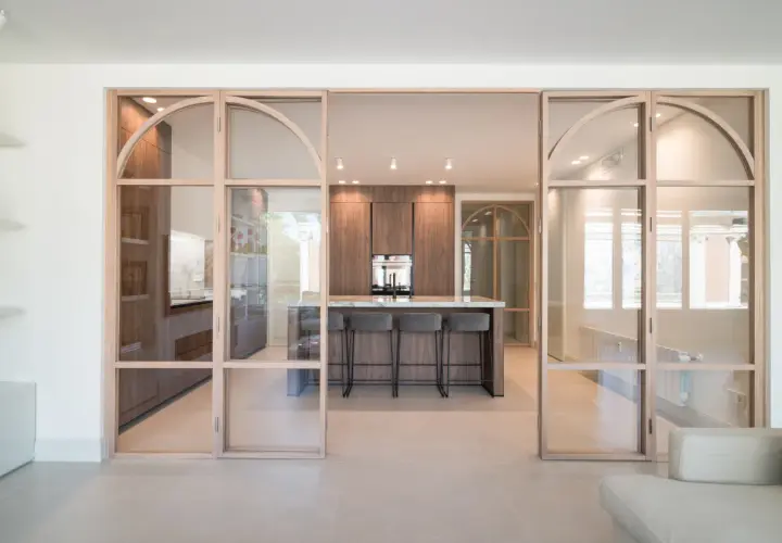 Moderna cocina semiabierta cristal con isla central y taburetes. Muebles de madera oscura y encimera de mármol.