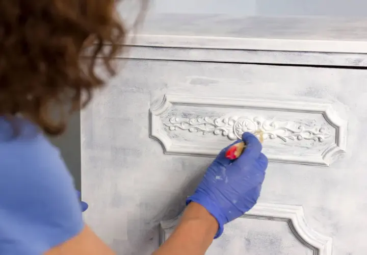 Persona con guantes azules aplica pintura blanca a un armario con detalles ornamentados, mostrando cómo pintar un armario sin lijar para un acabado desgastado.