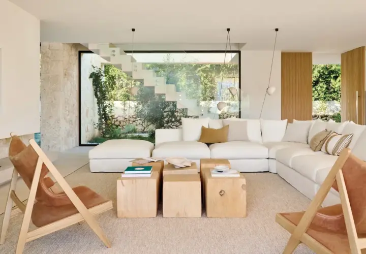 Amplio salón estilo mediterráneo con sofá blanco, mesas de madera y sillas de cuero, con vistas a un jardín exuberante a través de una escalera de cristal.