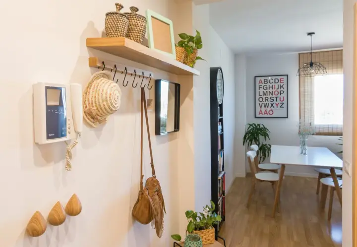 Pasillos con madera en la pared, un perchero con sombrero y bolso, y un intercomunicador. Al fondo, un comedor con mesa y sillas.