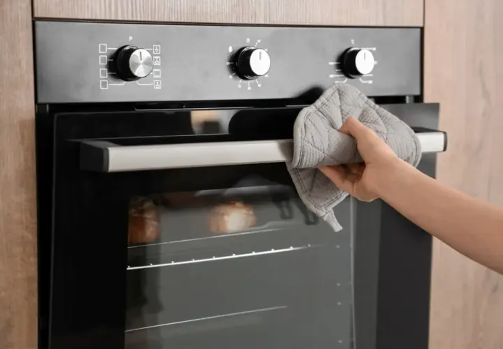 Mano con manopla sujetando la puerta del horno, donde se ve una sección de cable para horno y pan horneándose.