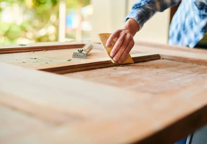 Mano lijando una puerta de madera exterior para restaurarla. Se ve el raspador al lado.