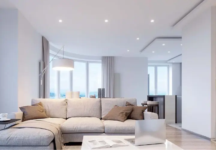 Salón moderno con sofá beige, mesa de centro y un techo pladur con focos empotrados. Ventanales curvos con vistas al mar.
