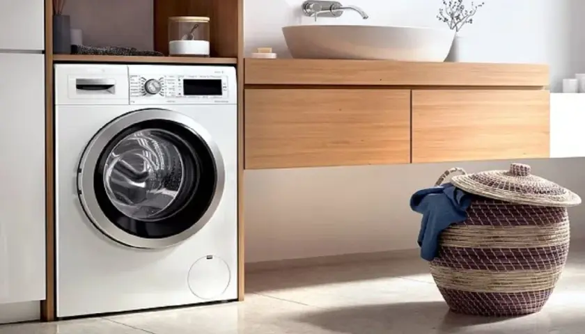 Lavadora moderna lista para instalar en un baño con cesta de ropa sucia.