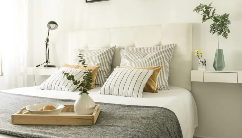 Ideas para como forrar un cabecero de cama: cama con cojines a rayas y lisos, bandeja con desayuno, lámpara y jarrón.