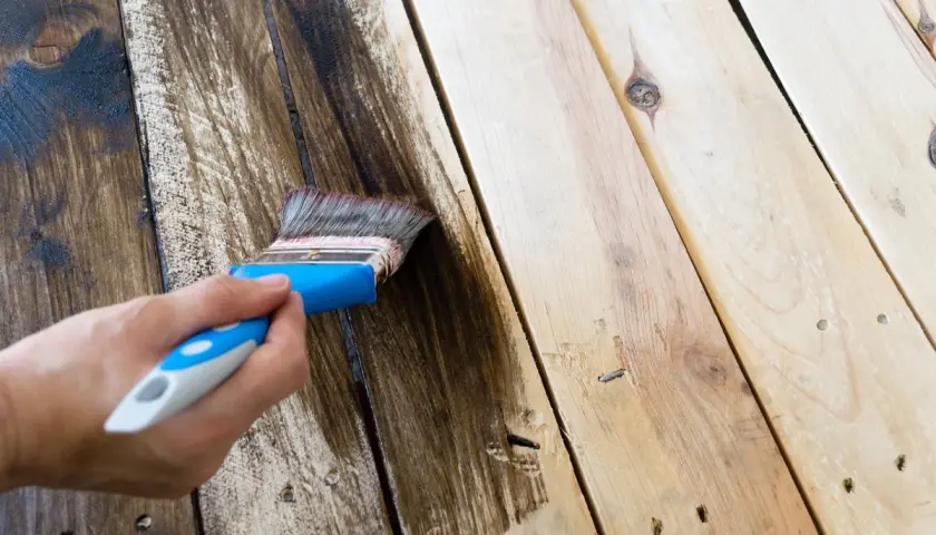 Mano aplicando tinte oscuro para pintar madera. El pincel deja un rastro de color sobre las tablas.