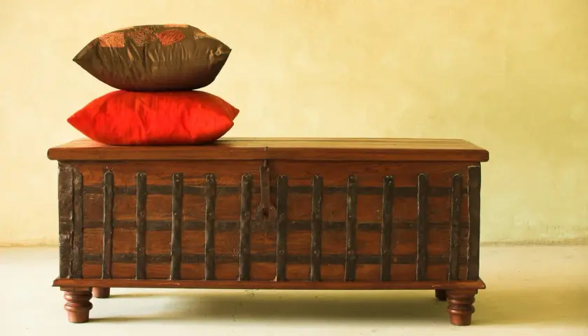 Un baúl antiguo de madera con detalles metálicos, coronado por dos cojines. Ideal para aprender como restaurar un baul antiguo de madera.