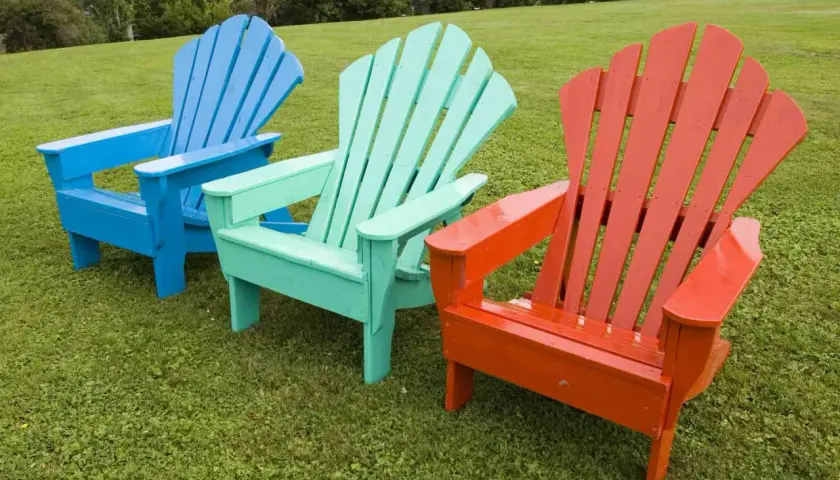 Tres sillas de madera estilo Adirondack, pintadas de azul, verde menta y naranja, lucen vibrantes sobre el césped. ¡Un proyecto de pintar sillas de madera sin lijar que da vida al exterior!