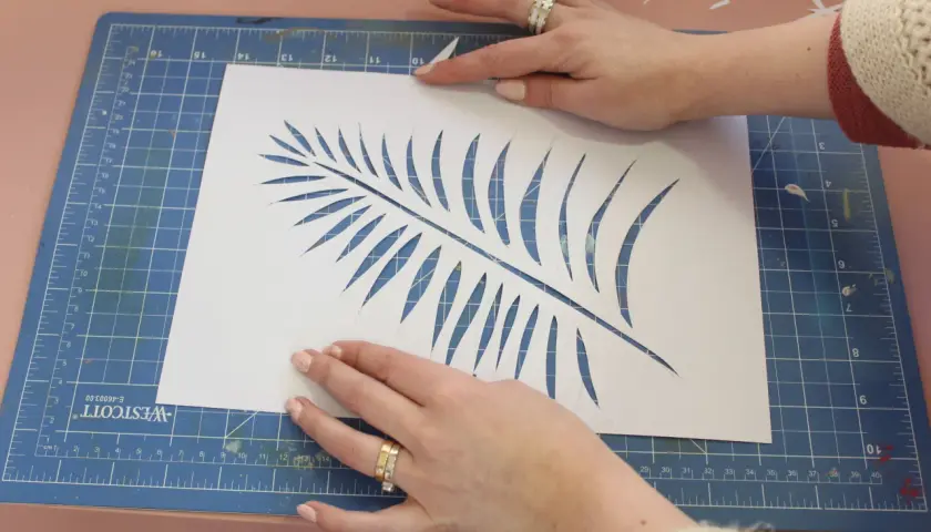 Manos cortando una hoja de helecho en papel blanco, un paso clave para saber como hacer plantillas para pintar.