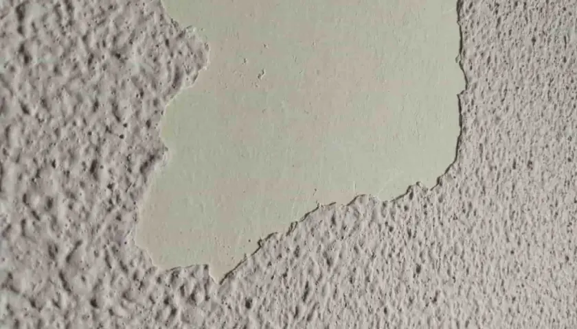 Detalle de pared con gotele plástico, donde una capa de pintura clara se desprende, revelando la textura rugosa debajo.