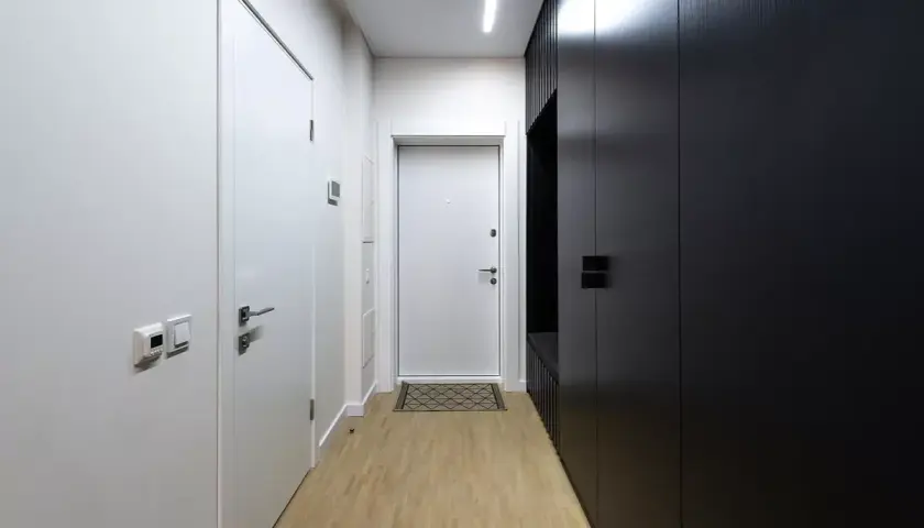 Pasillos estrechos y modernos con puertas blancas y un armario negro. El suelo de madera clara y la iluminación lineal crean un ambiente minimalista.