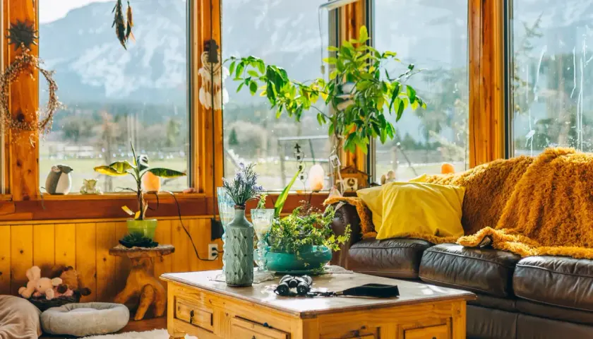 Un salón acogedor con sofá de cuero, manta naranja, cojín amarillo y mesa de centro de madera. Grandes ventanales con vistas a la montaña.