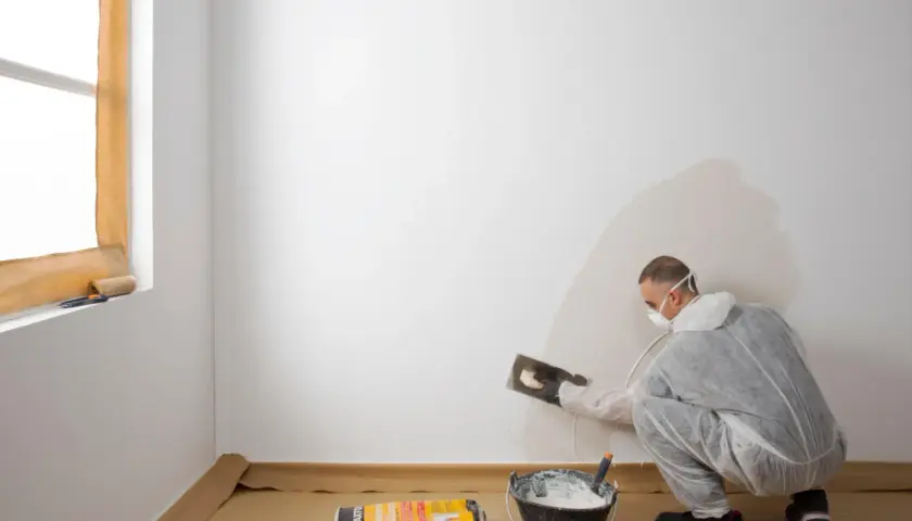 Hombre con mascarilla aplicando yeso para alisar paredes. Cubo con mezcla y bolsa de producto a su lado.