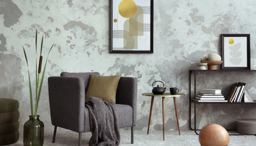 Salón moderno con pared texturizada que combina papel pintado y pintura. Sillón gris, mesa auxiliar dorada y estantería con arte abstracto.