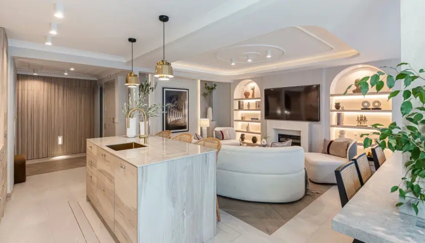 Cocina y sala de estar modernas con isla de mármol, sofás curvos y estanterías iluminadas. La iluminación ambiental crea una atmósfera acogedora, con suficientes **lúmenes por m2** para cada rincón.