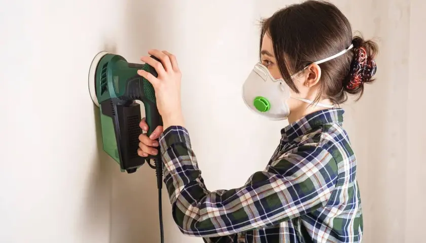 Mujer con mascarilla lijando paredes con una lijadora eléctrica.