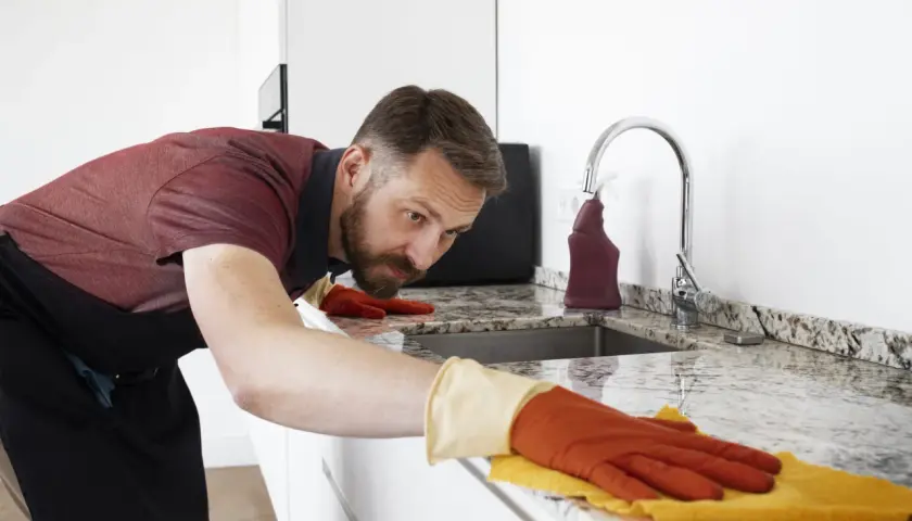 Hombre con guantes naranjas y amarillos, concentrado en limpiar encimera de granito con un paño.