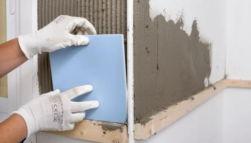Manos enguantadas colocando un azulejo azul sobre cemento fresco. Guía visual de como poner azulejos.