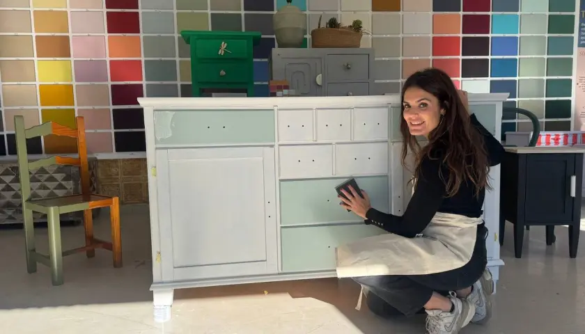 Mujer sonríe mientras lija un mueble de madera, mostrando cómo pintar un mueble de madera con un toque de color.