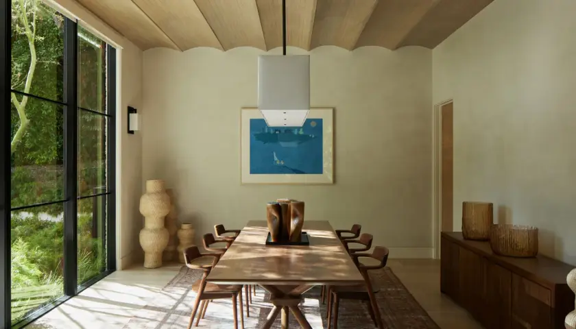 Comedor con mesa de madera, sillas de diseño y grandes ventanales. La decoración relajante se complementa con jarrones de mimbre y una pintura de paisaje marino.