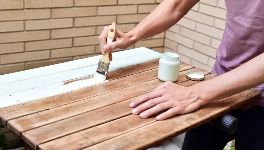 Persona pinta una mesa de madera barnizada con brocha y pintura blanca.