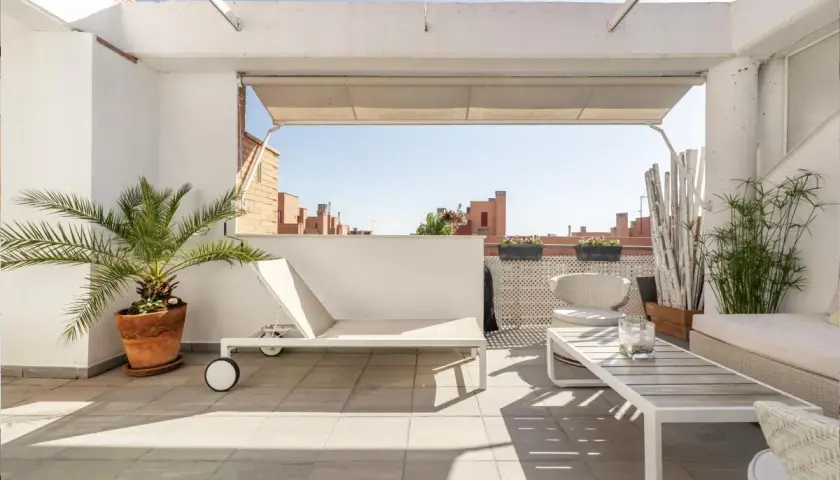 Ideas para decorar un ático: tumbona blanca, mesa de centro, sillas de mimbre y plantas verdes en una terraza soleada.