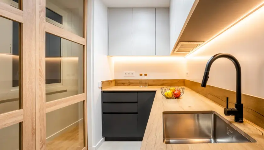 Cocina moderna con puertas de madera y cristal, encimera de madera, fregadero de acero inoxidable y grifo negro. Perfecta para reformas de cocinas pequeñas.