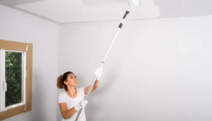 Mujer pintando el techo con rodillo. La pregunta de si que se pinta primero el techo o las paredes se resuelve pintando el techo.