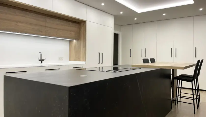 Cocina moderna con isla negra, encimeras blancas y armarios de madera. La iluminación LED bajo los armarios y en el techo resalta el diseño, mostrando cómo iluminar una cocina con estilo.