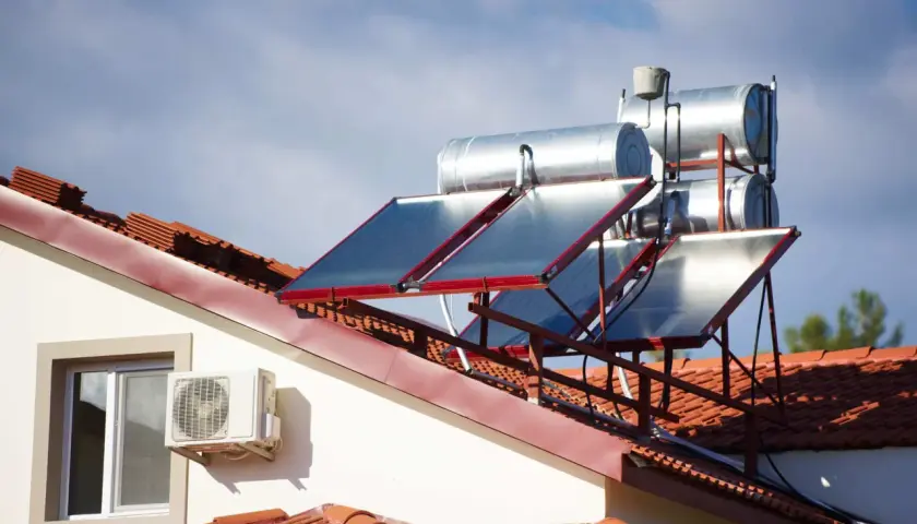 Paneles solares y tanques para agua caliente solar en un tejado de tejas rojas, bajo un cielo azul con nubes.
