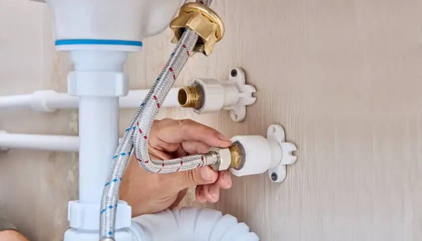 Mano ajustando la conexión de una manguera a la llave de paso de agua general, parte de la fontanería de un lavabo.