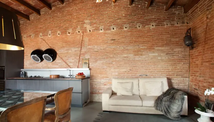 Un salón moderno con una pared rústica interior de ladrillo visto, sofá beige, cocina integrada y sillas de cuero.