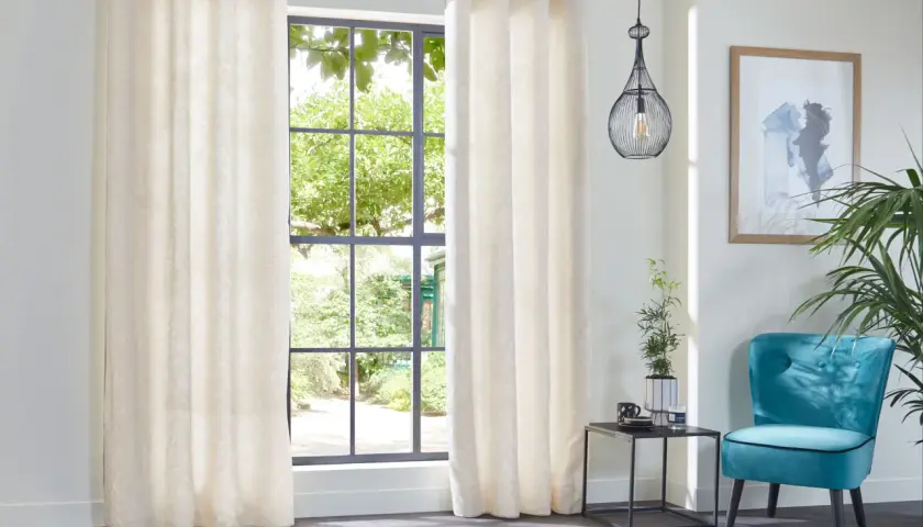 Elegantes cortinas de tela crema con sutil estampado, ideales para distintos tipos de cortinas para ventanas, complementan un sillón turquesa y un cuadro abstracto.