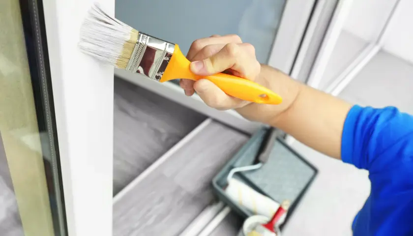 Mano pintando un marco de ventana blanco con una brocha. Se ve una bandeja de pintura y un rodillo en el fondo. Ideal para pintura para pintar plástico.