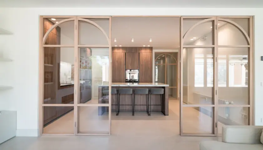 Moderna cocina semiabierta cristal con isla central y taburetes. Muebles de madera oscura y encimera de mármol.