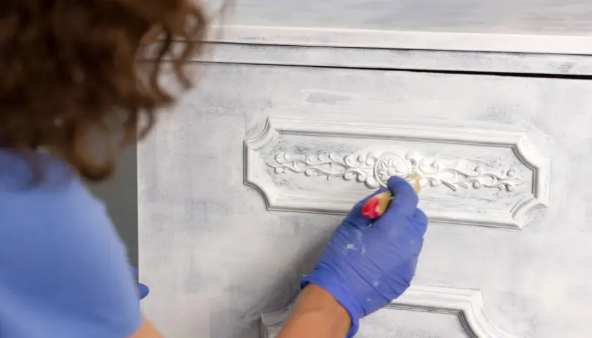 Persona con guantes azules aplica pintura blanca a un armario con detalles ornamentados, mostrando cómo pintar un armario sin lijar para un acabado desgastado.