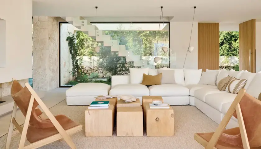 Amplio salón estilo mediterráneo con sofá blanco, mesas de madera y sillas de cuero, con vistas a un jardín exuberante a través de una escalera de cristal.