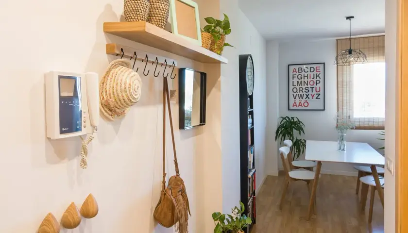 Pasillos con madera en la pared, un perchero con sombrero y bolso, y un intercomunicador. Al fondo, un comedor con mesa y sillas.