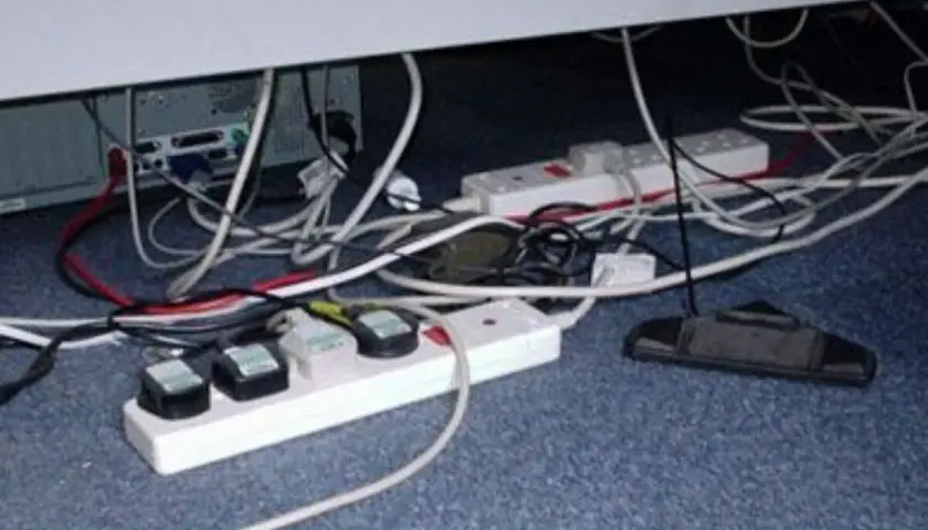 Un enredo de cables y regletas bajo un escritorio. Un ejemplo de qué es un contacto eléctrico indirecto, con cables expuestos y enchufes.