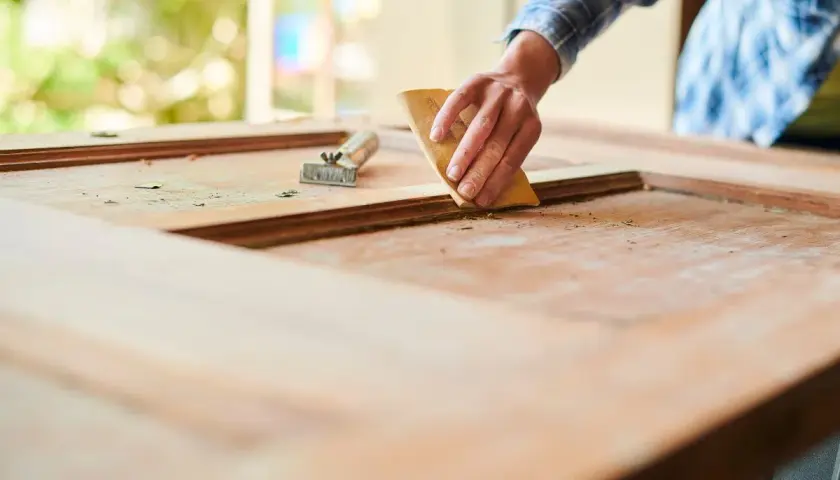 Mano lijando una puerta de madera exterior para restaurarla. Se ve el raspador al lado.
