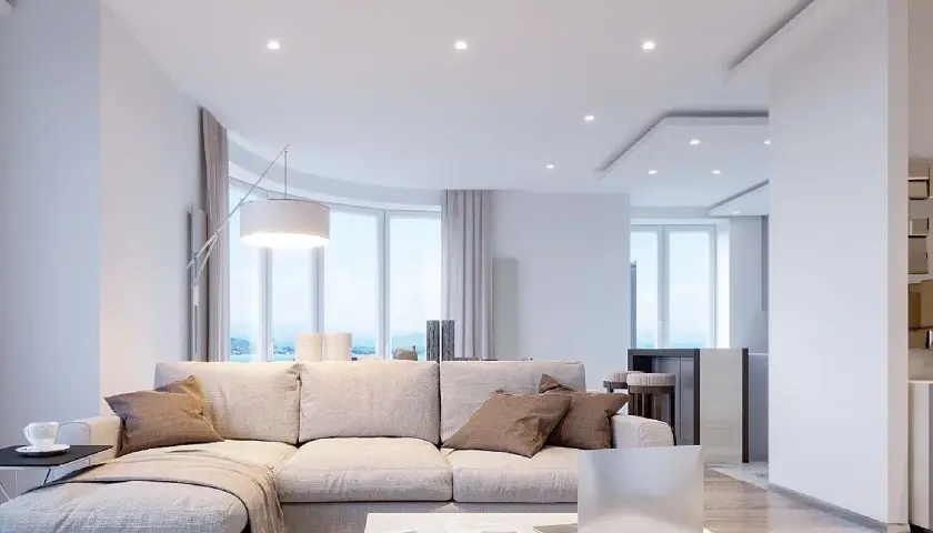 Salón moderno con sofá beige, mesa de centro y un techo pladur con focos empotrados. Ventanales curvos con vistas al mar.