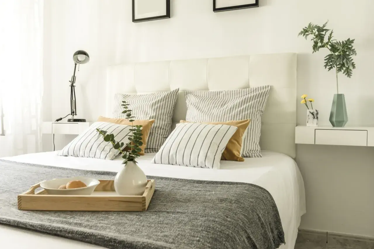 Ideas para como forrar un cabecero de cama: cama con cojines a rayas y lisos, bandeja con desayuno, lámpara y jarrón.