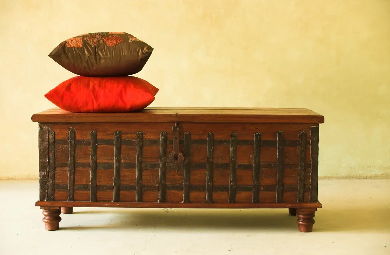 Un baúl antiguo de madera con detalles metálicos, coronado por dos cojines. Ideal para aprender como restaurar un baul antiguo de madera.