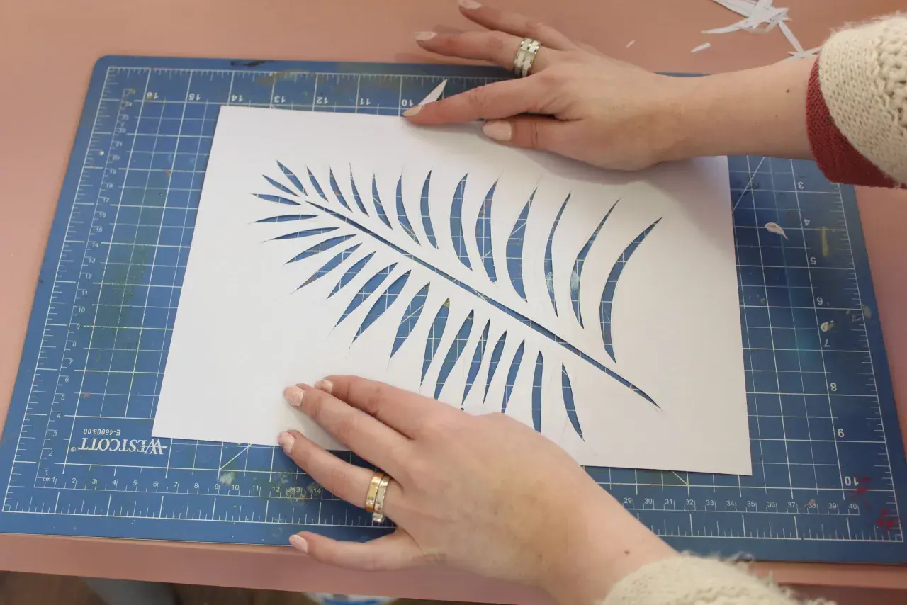 Manos cortando una hoja de helecho en papel blanco, un paso clave para saber como hacer plantillas para pintar.