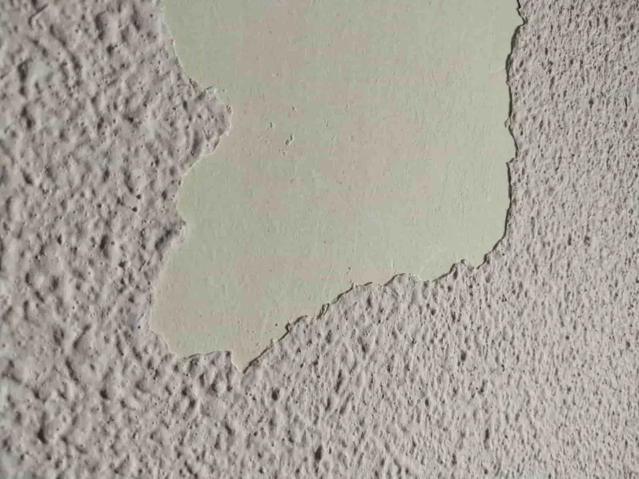 Detalle de pared con gotele plástico, donde una capa de pintura clara se desprende, revelando la textura rugosa debajo.